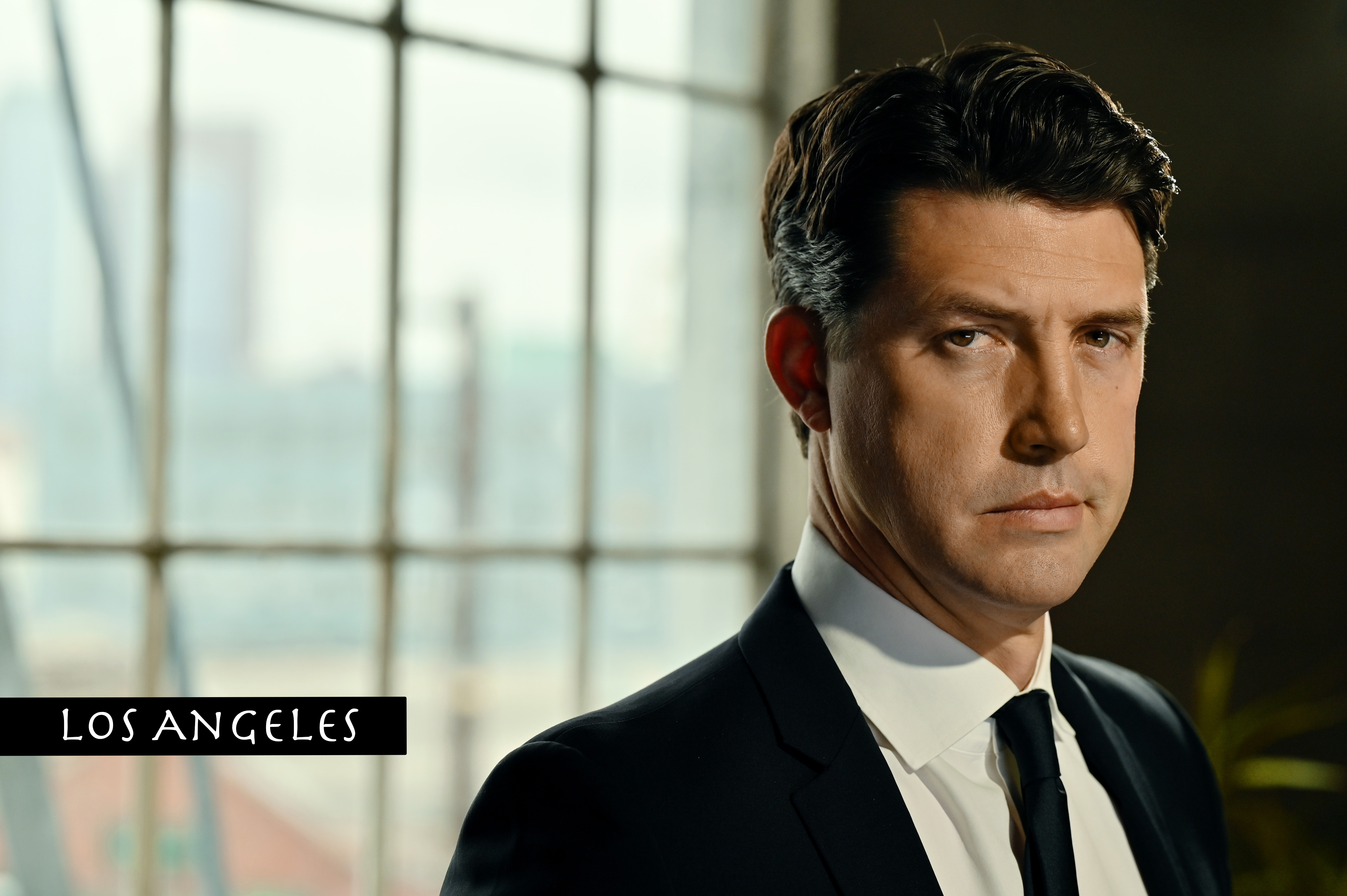 Tug Coker in a suit standing with a large window behind him.